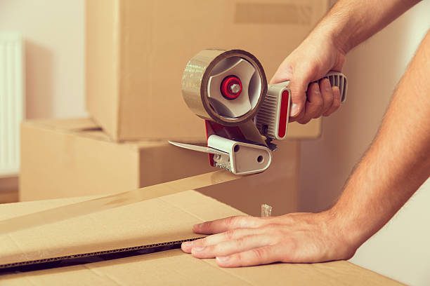 Close up of a guy's hands holding packing machine and sealing cardboard boxes with duct tape